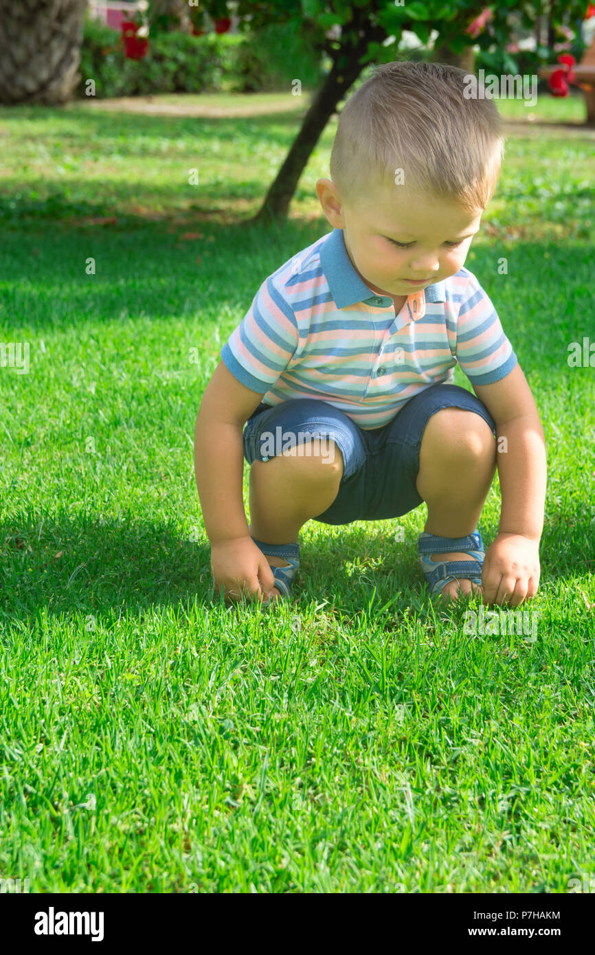 Carino bionda caucasica Baby Boy con gli occhi blu bimbo di 2 anni si siede su Haunches su erba verde nel parco della città durante la riproduzione. Vivid luminosa giornata estiva Gree Foto Stock