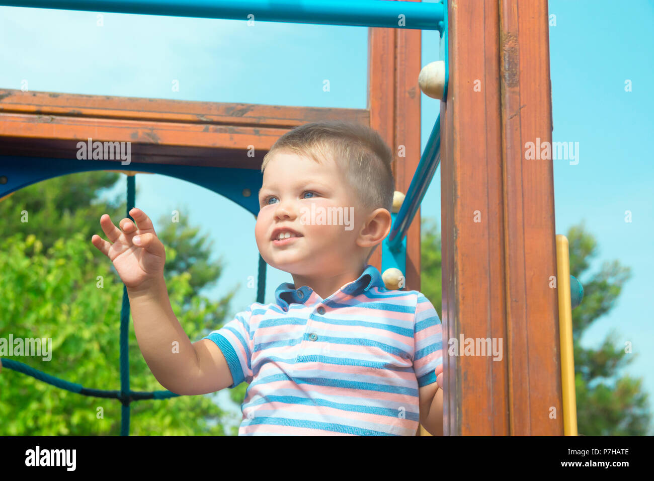 Carino bionda caucasica Baby Boy con gli occhi blu bimbo di 2 anni si erge sul parco giochi nel parco cittadino sorridente. Vivid luminosa giornata estiva fogliame verde B Foto Stock