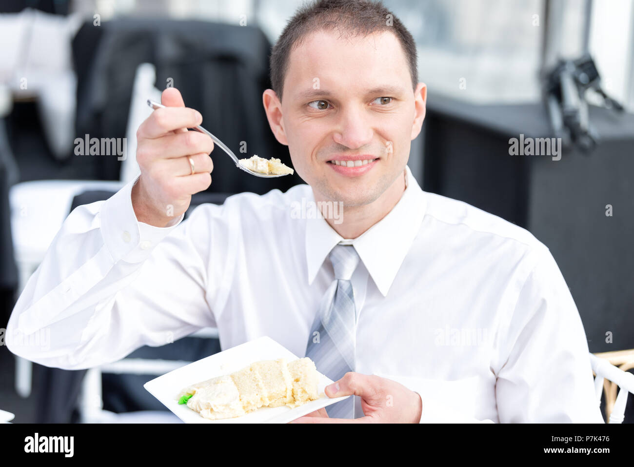 Giovane uomo felice in business abito formale shirt sorridente di mangiare la torta con la forcella a ricevimento di nozze cena dessert seduti a tavola Foto Stock
