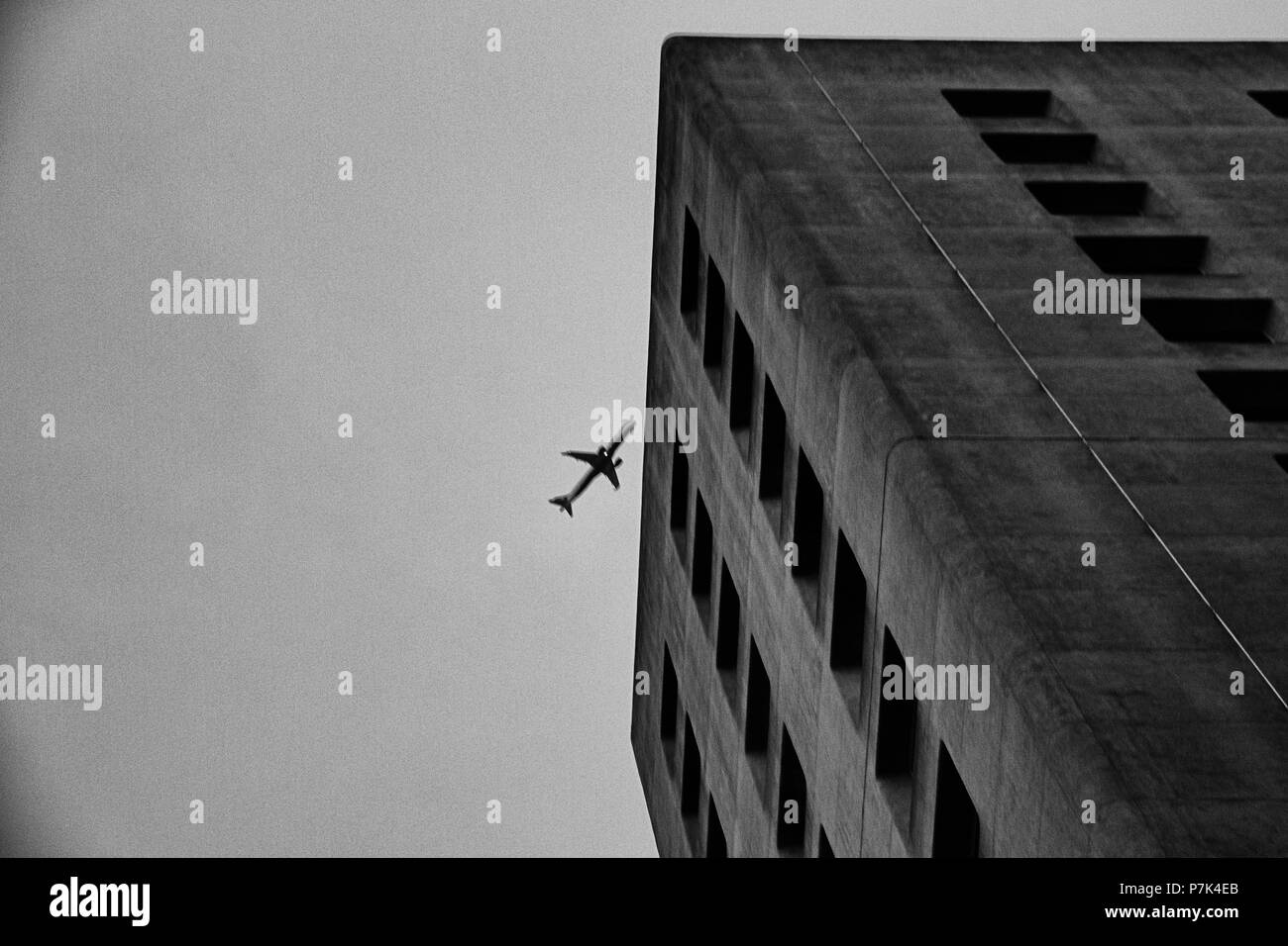 Immagine in bianco e nero di un calcestruzzo edificio per uffici con un aereo nel cielo che sembra quasi di schiantarsi su di esso. Foto Stock