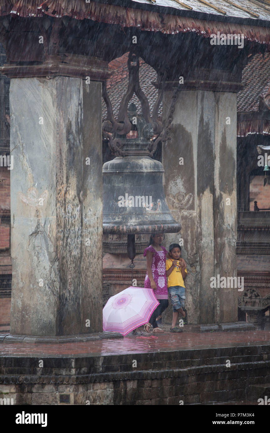 Donna locale con bambini in cerca di protezione dalla pioggia sotto una campana, tempio, Kathmandu, Nepal Foto Stock