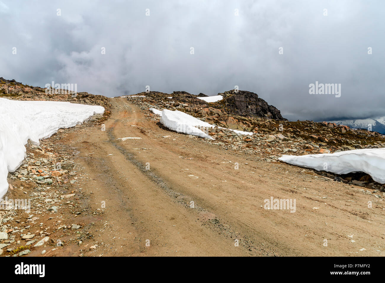 Strada sterrata, rocky strada verso la cima di una montagna rocciosa coperta con cumuli di neve, una nebbiosa blue sky in background con nuvole bianche su un giorno di estate Foto Stock
