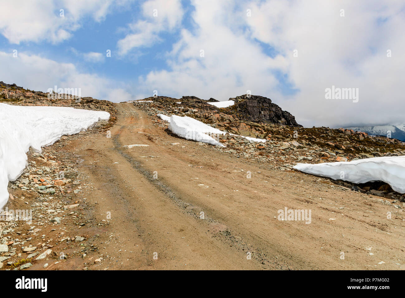 Strada sterrata, rocky strada verso la cima di una montagna rocciosa coperta con cumuli di neve, una nebbiosa blue sky in background con nuvole bianche su un giorno di estate Foto Stock