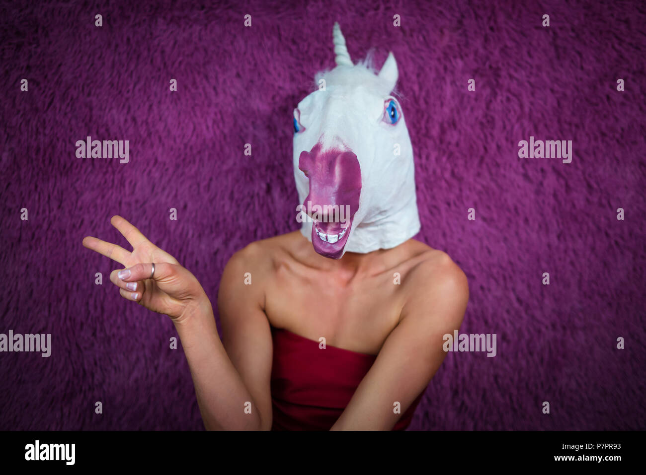 Funny Girl unicorn flirta e gesticulating. Freaky giovane donna in maschera comica sorge su sfondo viola. Ritratto di signora inusuale in abito rosso. Foto Stock
