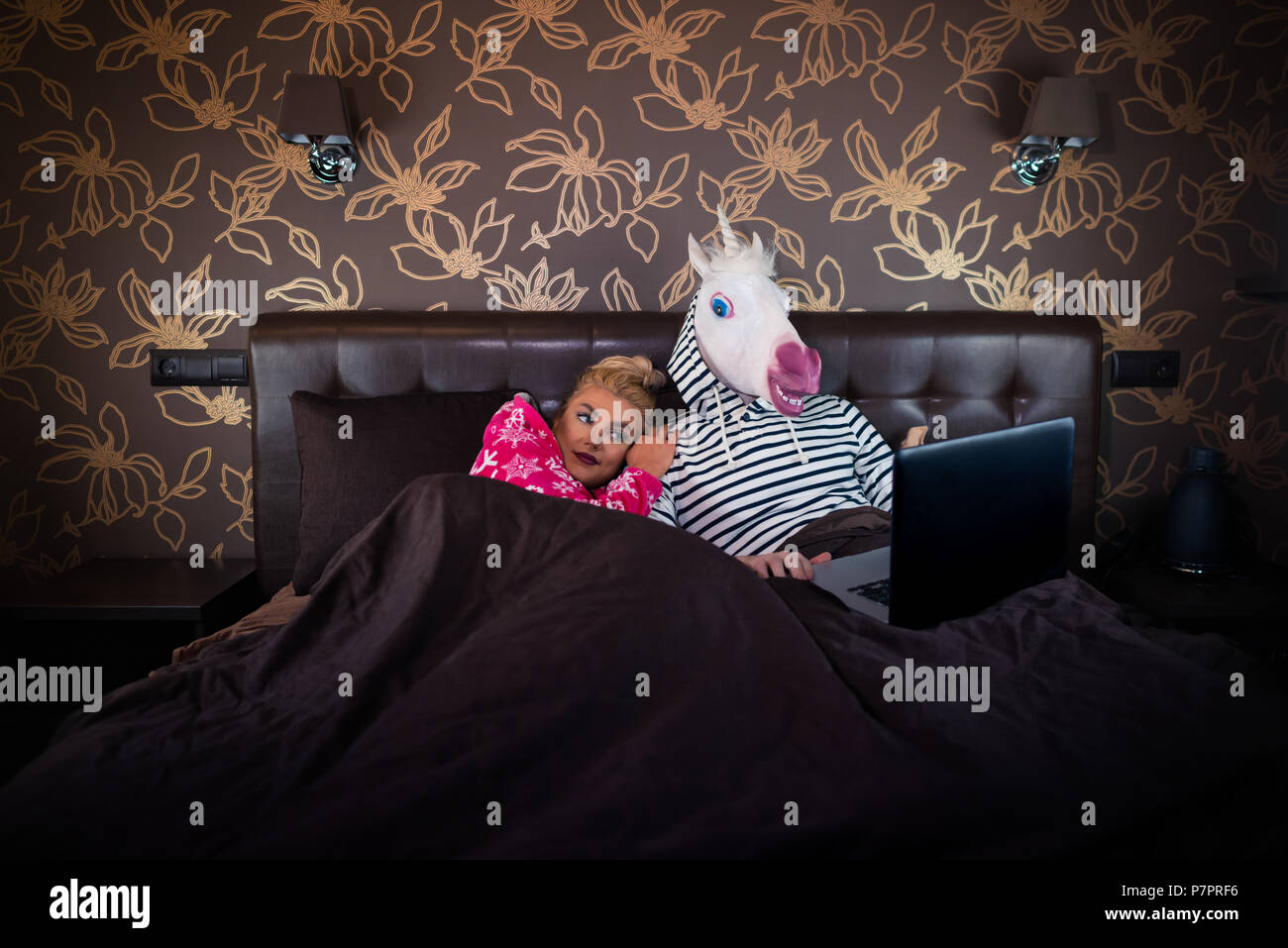 Bella ragazza è sdraiato sul letto con divertenti fidanzato in maschera comica. Coppia insolita è la visione di film sul computer portatile presso l'elegante camera da letto. Foto Stock