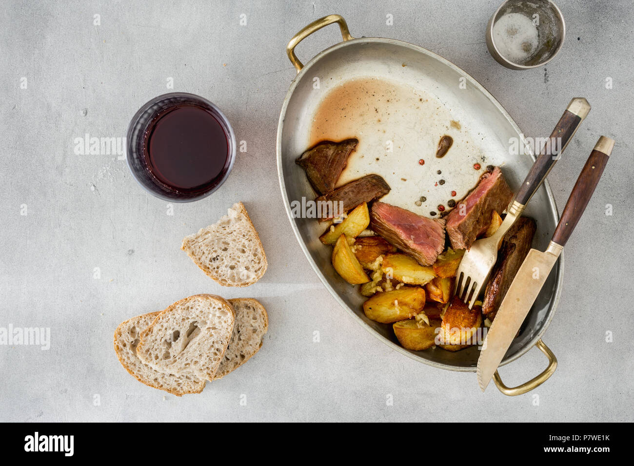 Vista da sopra le fette di carne di manzo con patate in padella sulla superficie della luce con un bicchiere di vino rosso Foto Stock