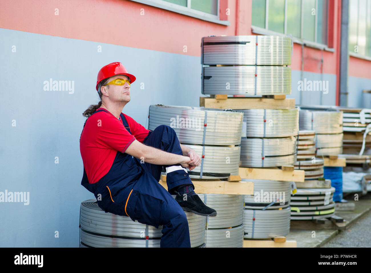 Operaio di fabbrica con casco rosso in appoggio al di fuori di una fabbrica Foto Stock