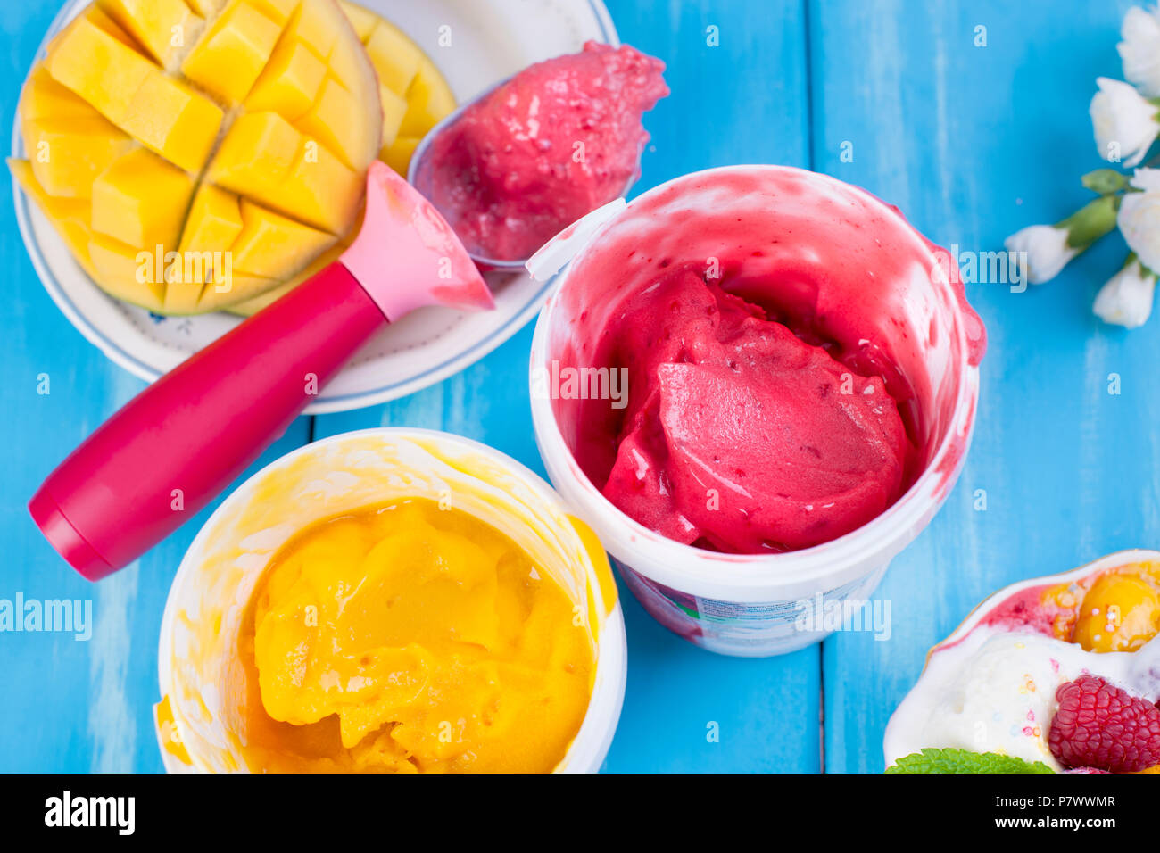Gelato di frutta con mango e lamponi in una benna pack. Cucchiaio per gelato. Sfondo blu e fiori. copia di spazio. Foto Stock
