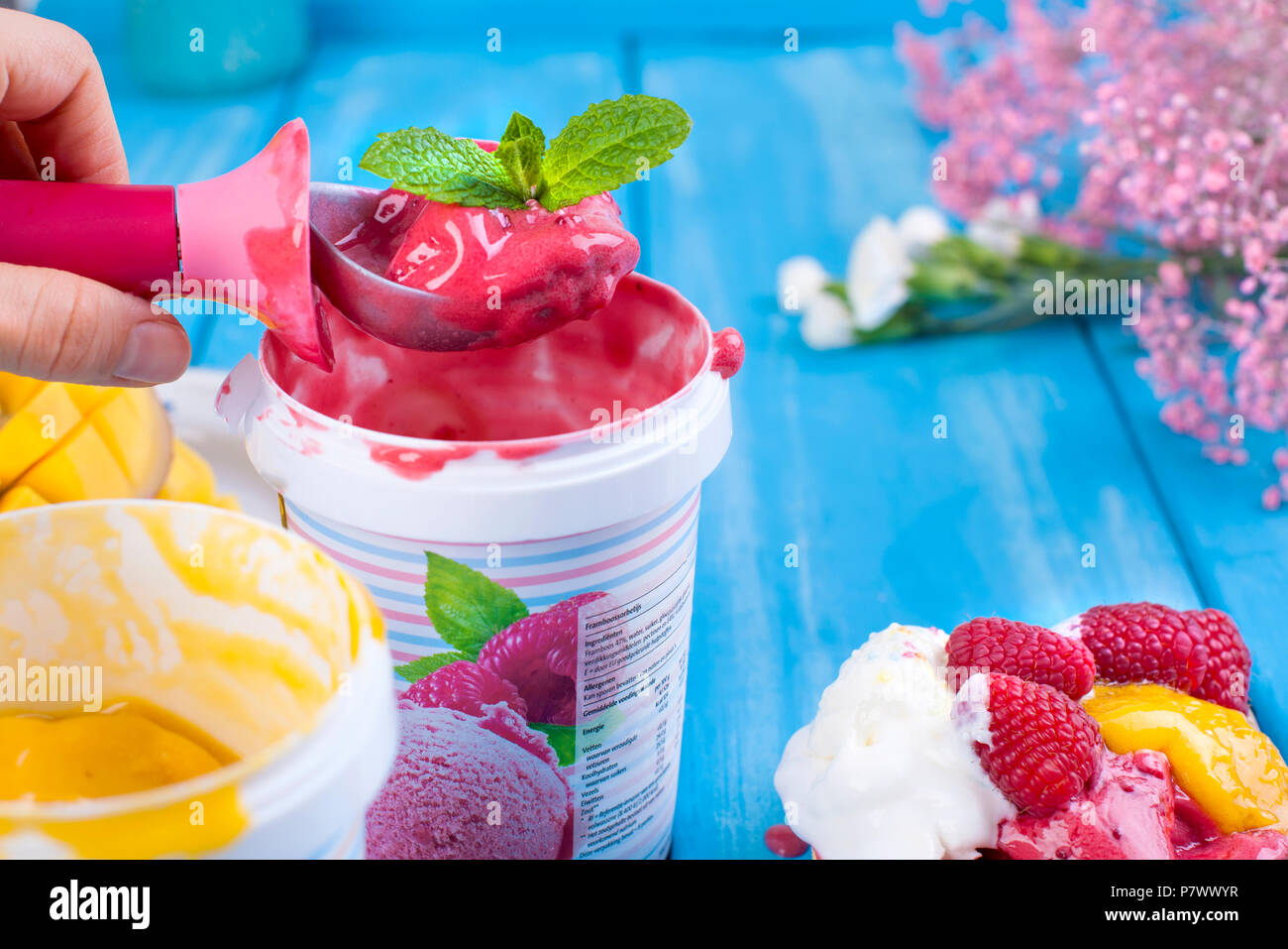 Gelato di frutta con mango e lamponi in una benna pack. Cucchiaio per gelato in mano. Sfondo blu e fiori. Card Foto Stock
