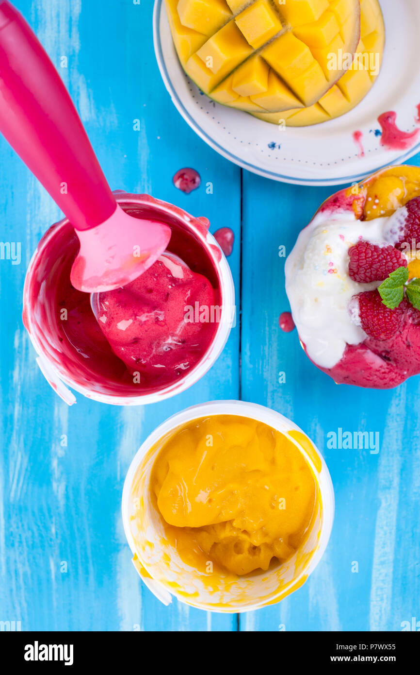 Gelato di frutta con mango e lamponi in una benna pack. Cucchiaio per gelato. Sfondo blu e fiori. Dolce con Foto Stock