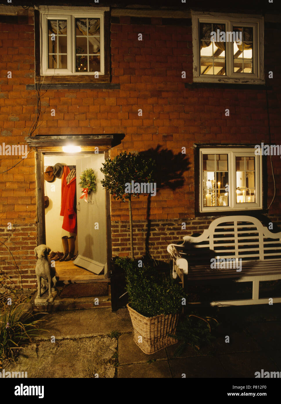 Bay Tree in pentola accanto al banco di Lutyens al di fuori del cottage di notte con finestre illuminate Foto Stock