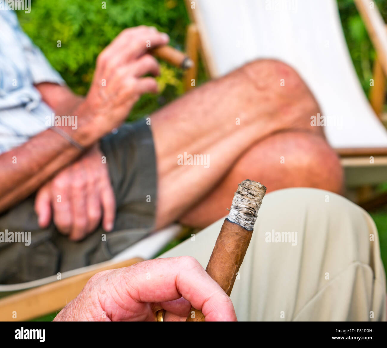 In prossimità di due alti uomini seduti in sedie a sdraio nel giardino estivo godendo di fumare un sigaro cubano, London, England, Regno Unito Foto Stock