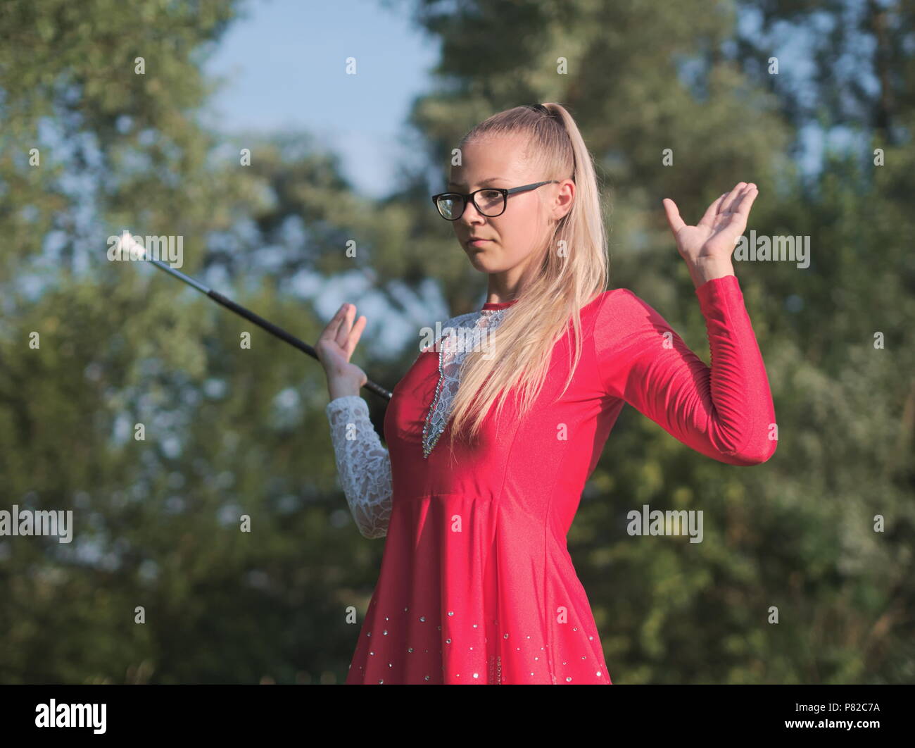 Bespectacled Blonde Teen Majorette ragazza fa roteare Baton all aperto in abito rosso Foto Stock