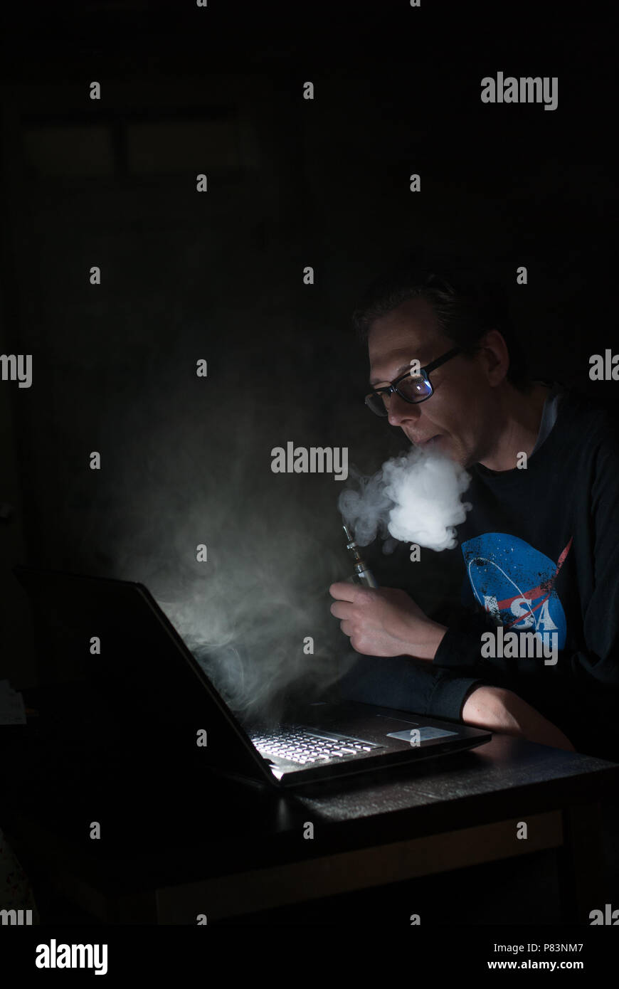 Programmatore pagine lavora a un computer portatile mentre vaping in una stanza buia. Foto Stock