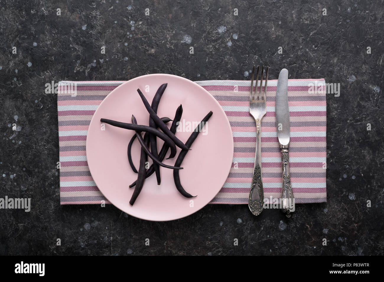 Stringa di viola gli asparagi Fagioli baccelli sulla piastra di rosa con posate su dark grunge tabella. La dieta, mangiare sano concetto. Vista dall'alto. Foto Stock