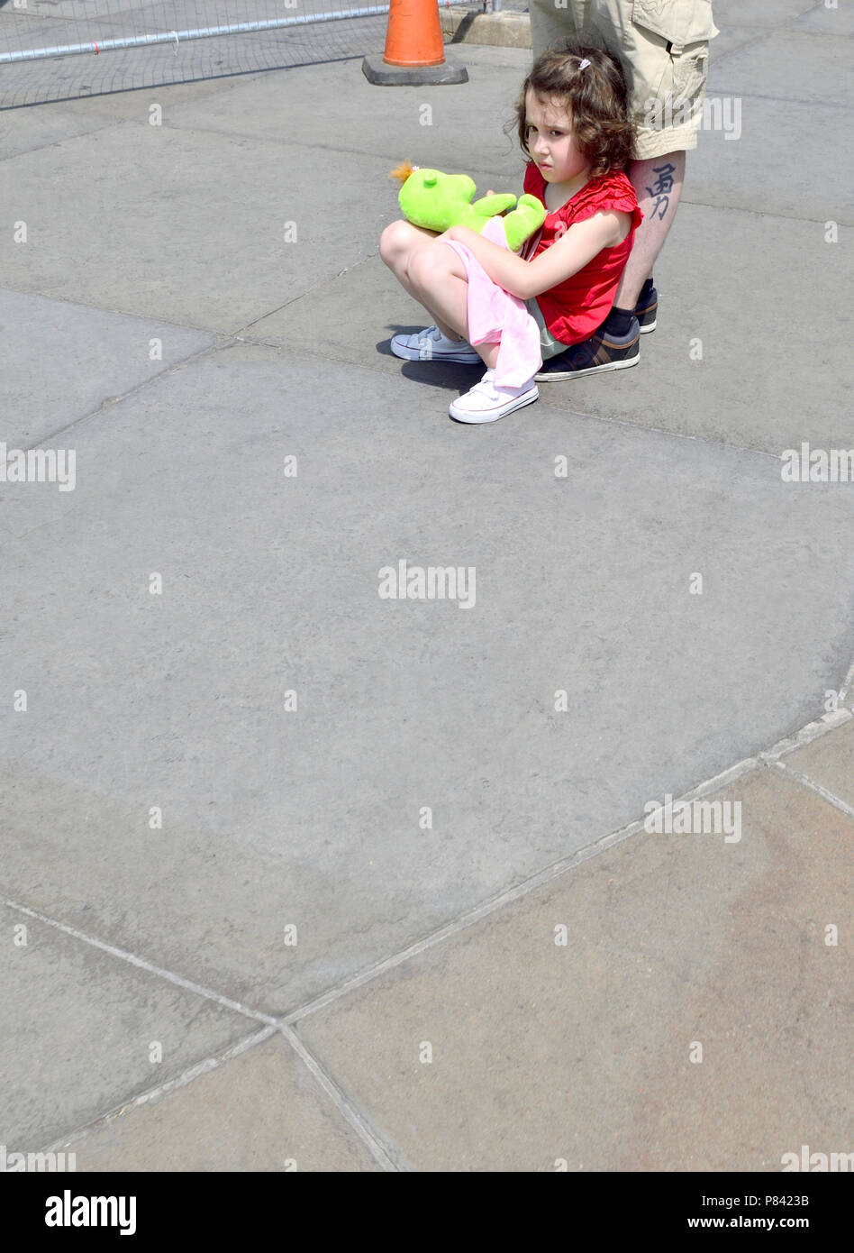 Giovane ragazza seduta con un giocattolo morbido in Trafalgar Square a Londra, Inghilterra, Regno Unito. Appoggiata al suo padre gamba tatuato Foto Stock