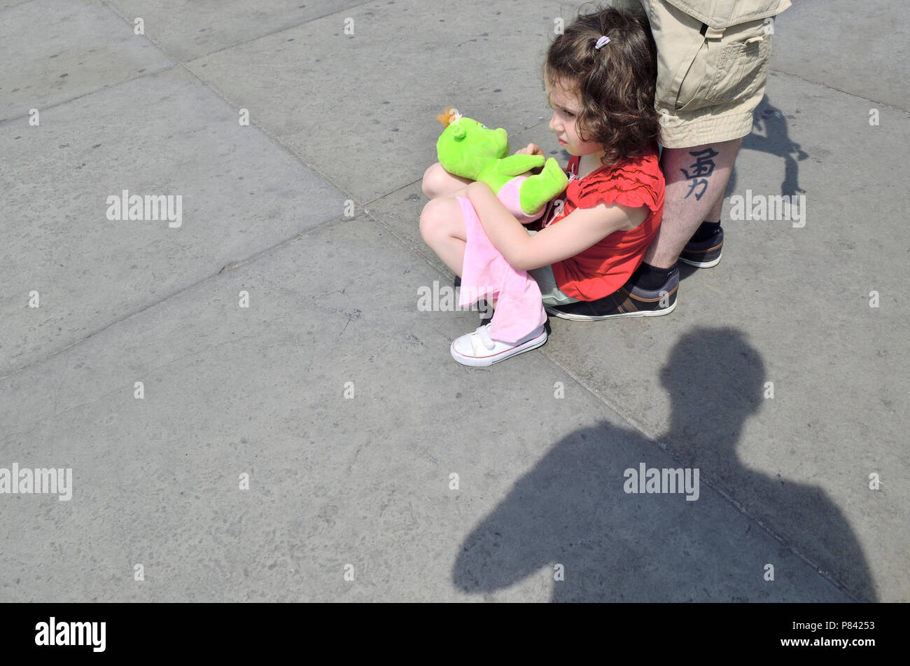 Giovane ragazza seduta con un giocattolo morbido in Trafalgar Square a Londra, Inghilterra, Regno Unito. Appoggiata al suo padre gamba tatuato Foto Stock
