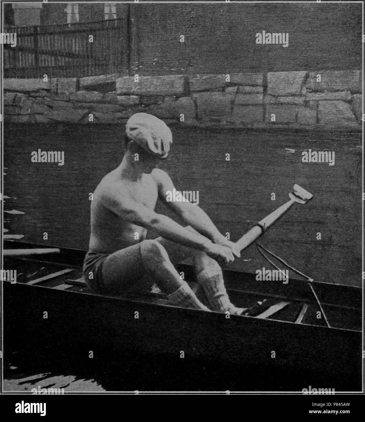 Fotografia in bianco e nero di un uomo che indossa un cappello, corti e calze, ma nessuna camicia, seduti in un scull e canottaggio un unico remo con entrambe le mani, con un terrapieno di pietra in background, 1906. La cortesia Internet Archive. () Foto Stock