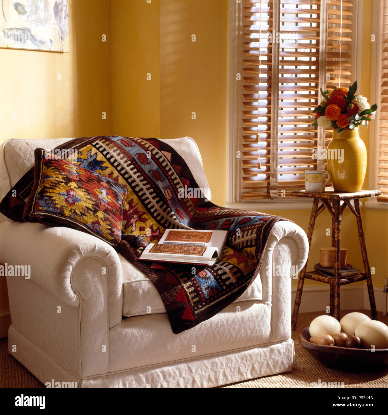 Blu, rosso e giallo arazzo buttare su una poltrona bianca accanto ad una piccola tabella di bambù nella parte anteriore della finestra con persiane piantagioni Foto Stock