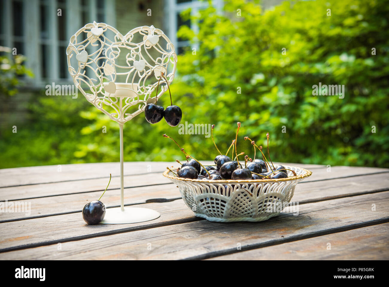 Fresche ciliege rosse bacche in una piastra bianca sul tavolo di legno con candelabro a forma di cuore all'aperto nel giardino. Un posto romantico. Foto Stock