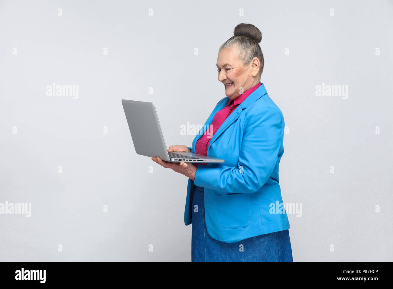 Età della donna in piedi e tenendo il laptop e guardando lo schermo con faccia soddisfatta. Nonna in azzurro tuta e raccolti i capelli grigi bun hairstyle Foto Stock