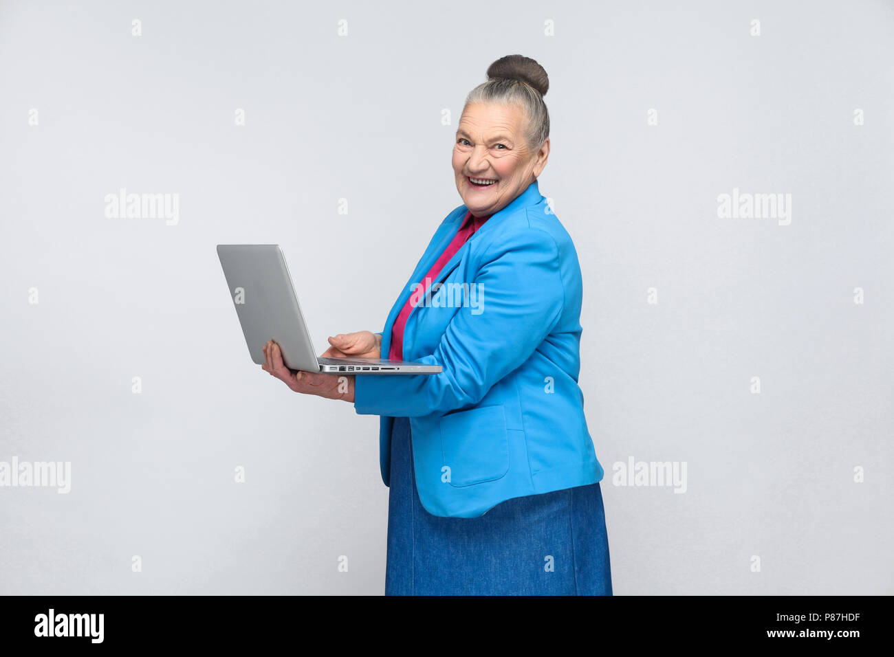 Età della donna in piedi e tenendo il laptop e guardando la fotocamera con faccina sorridente. Nonna in azzurro tuta e raccolti i capelli grigi bun hairstyle. i Foto Stock