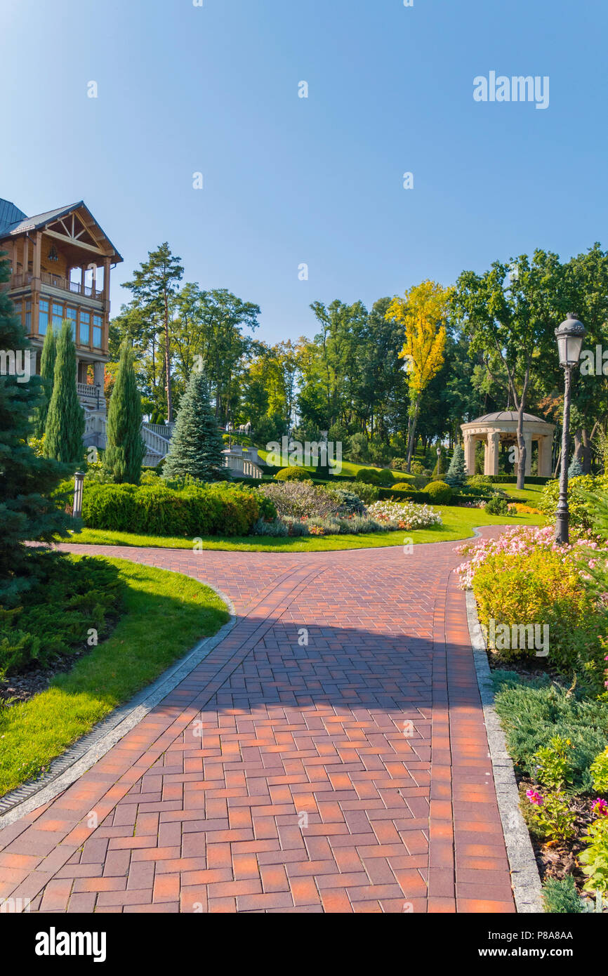 Un bellissimo vicolo lastricato nel verde di un parco con alberi ad alto fusto, fiori e lanterne, disteso di fronte a un enorme country house, una dimora. riposo-plac Foto Stock