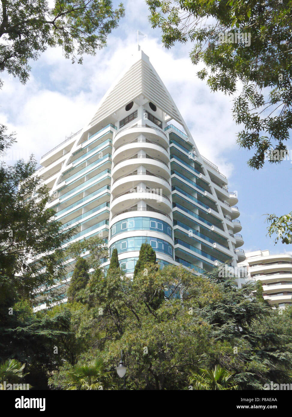 Al di sopra del alberi verdi e alberi di palma contro il blu cielo nuvoloso torri un alto edificio bianco di un edificio residenziale o hotel con grande balco blu Foto Stock