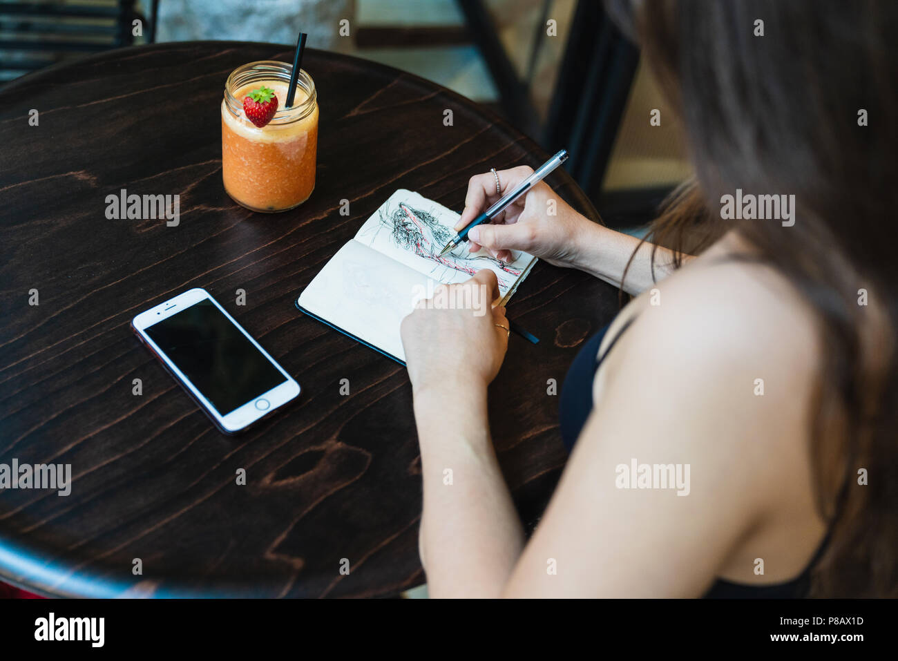 Da sopra il colpo di anonimo giovane donna seduta al tavolo del bar e fare schizzi in notebook di piccole dimensioni Foto Stock