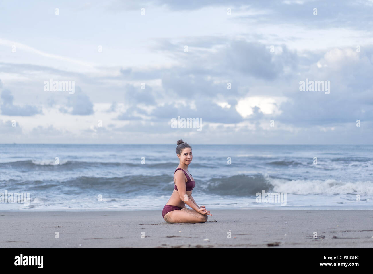 Vista laterale della bella giovane donna a praticare yoga in lotus pongono (padmasana) con gyan mudra sulla riva del mare e guardando la fotocamera Foto Stock