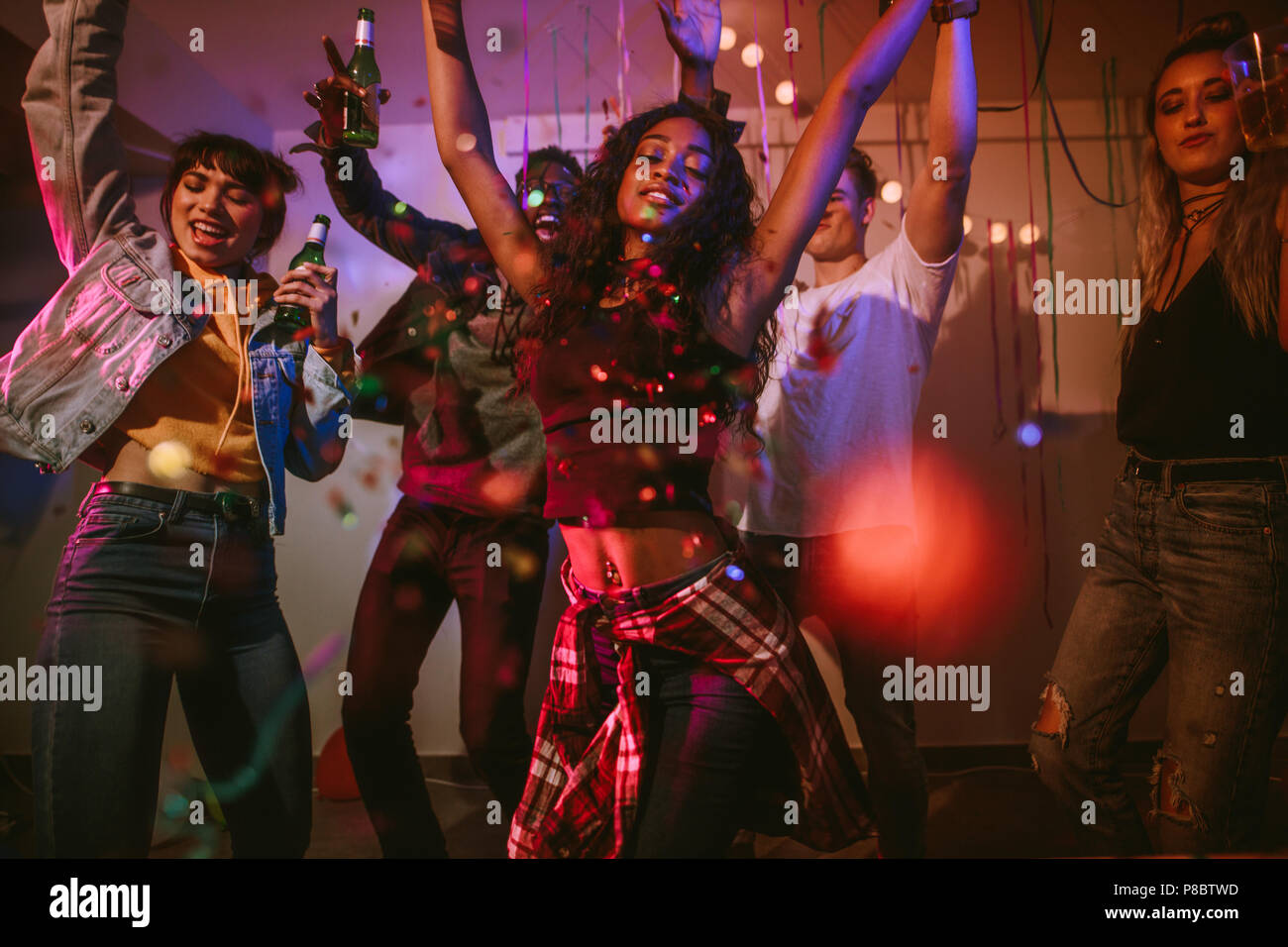 Giovani uomini e donne aventi il divertimento a un colorato house party con i confetti tutto intorno. Amici ballare e festeggiare azienda bevande a house party. Foto Stock