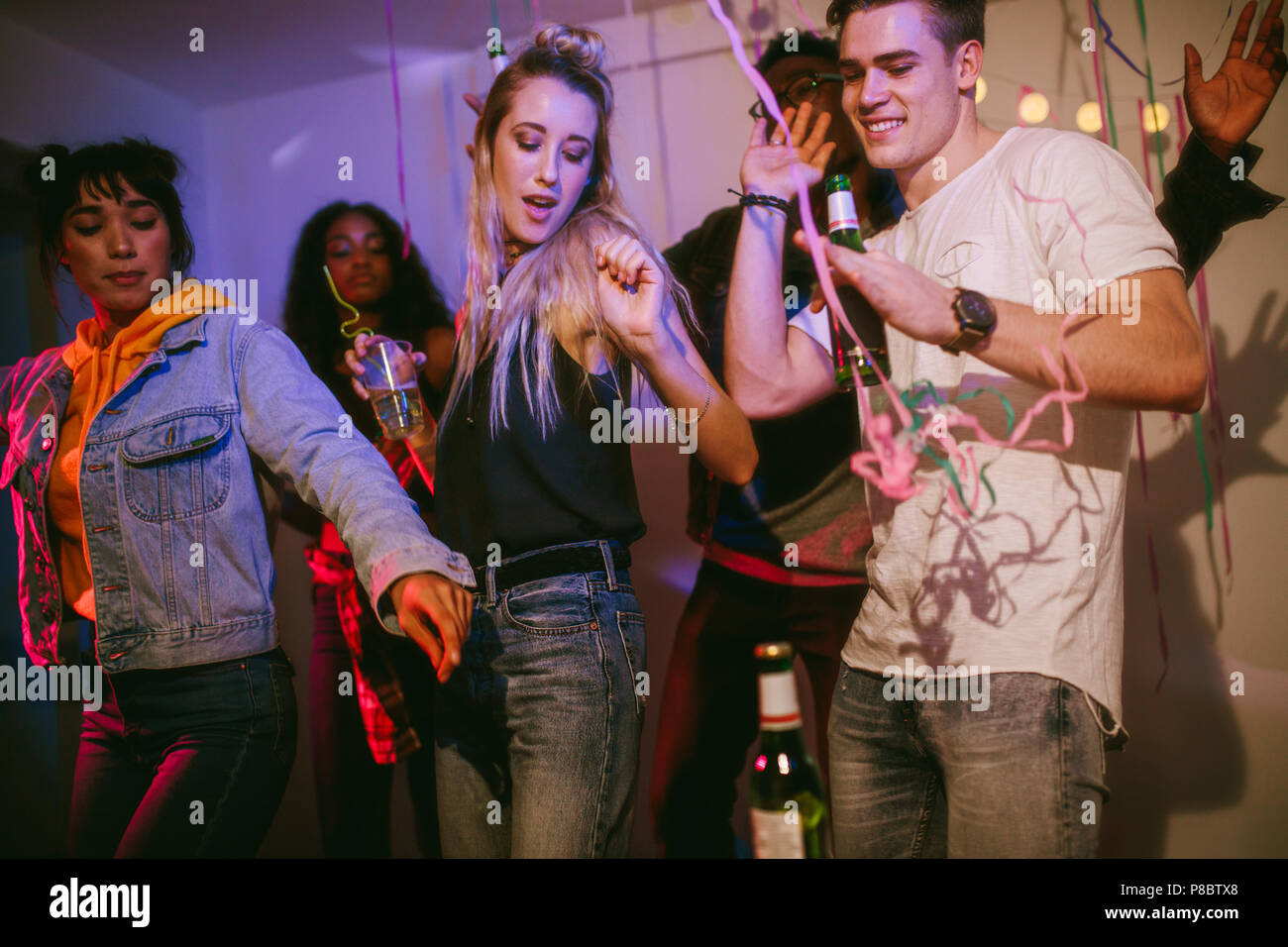 Amici ballare tenendo bevande e divertirsi a una festa. Giovani uomini e donne godendo di un colorato house party con nastro coriandoli tutti intorno a Foto Stock