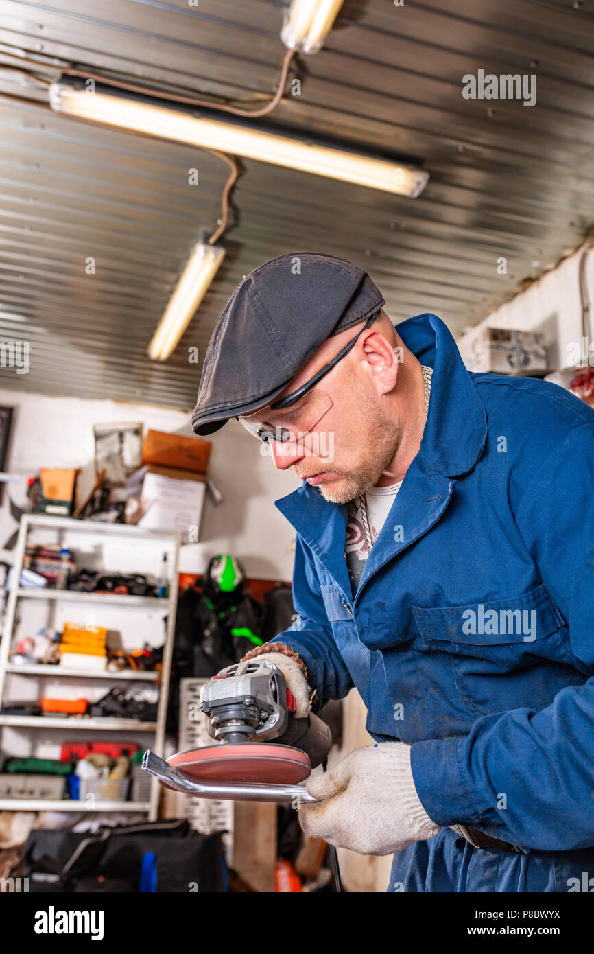 Un giovane uomo saldatore in una blu T-shirt, occhiali di protezione e guanti di costruzione metallica elabora una smerigliatrice angolare in garage, sullo sfondo di un sacco di strumenti Foto Stock