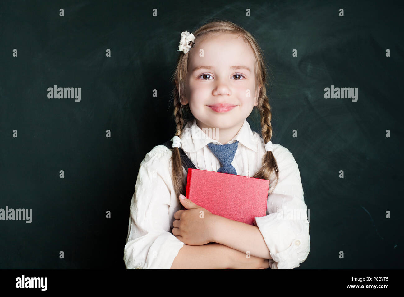 Carino Little Schoolgirl bambino con la scuola prenota sulla lavagna verde dello sfondo. Pupilla sorridente in aula (bambino 5-6 anni) Foto Stock