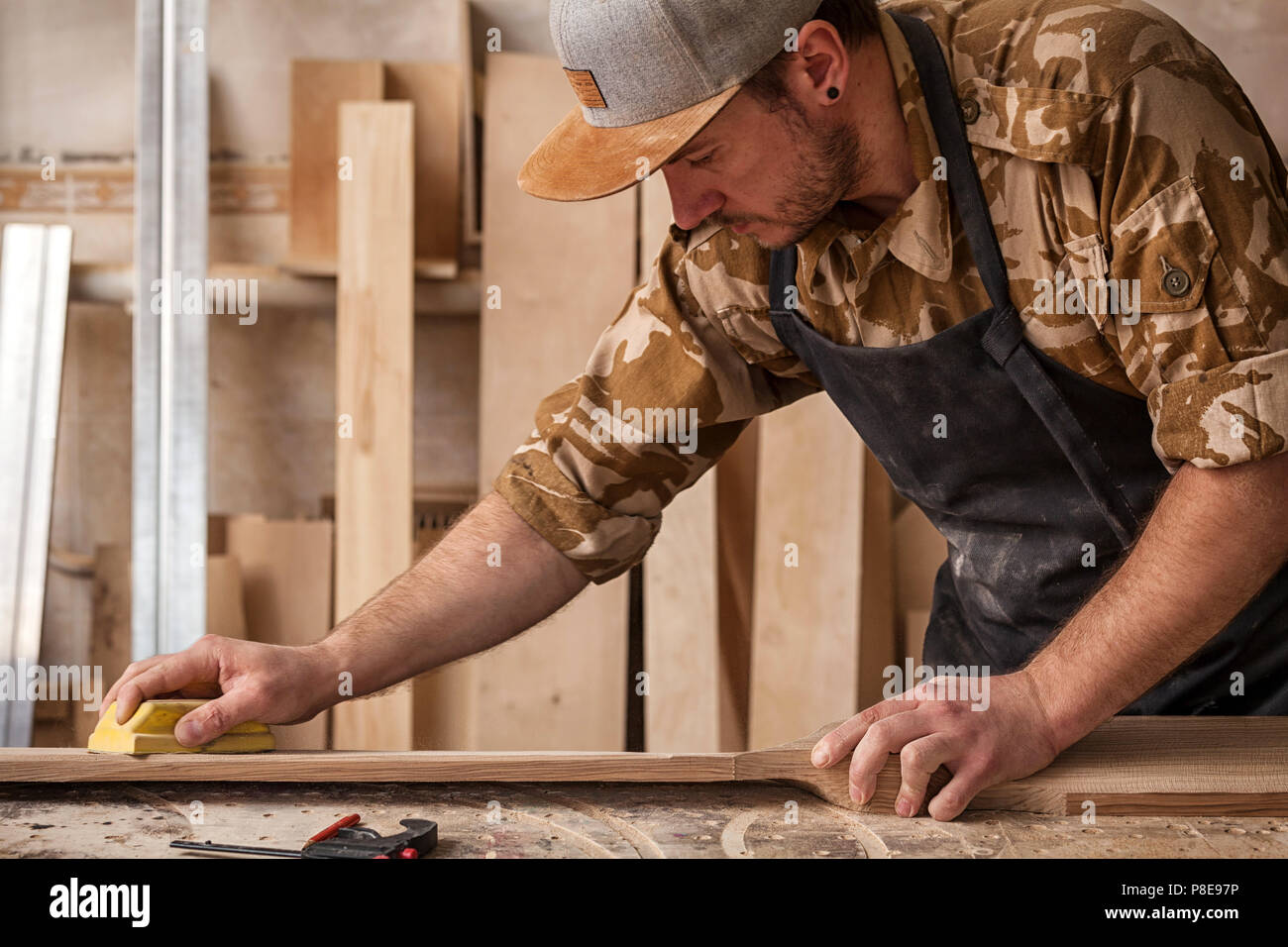 Un uomo di lavoro in un cappuccio e camicia lucida il blocco di legno con carta vetrata prima della verniciatura in officina, in background, strumenti e mach di perforazione Foto Stock