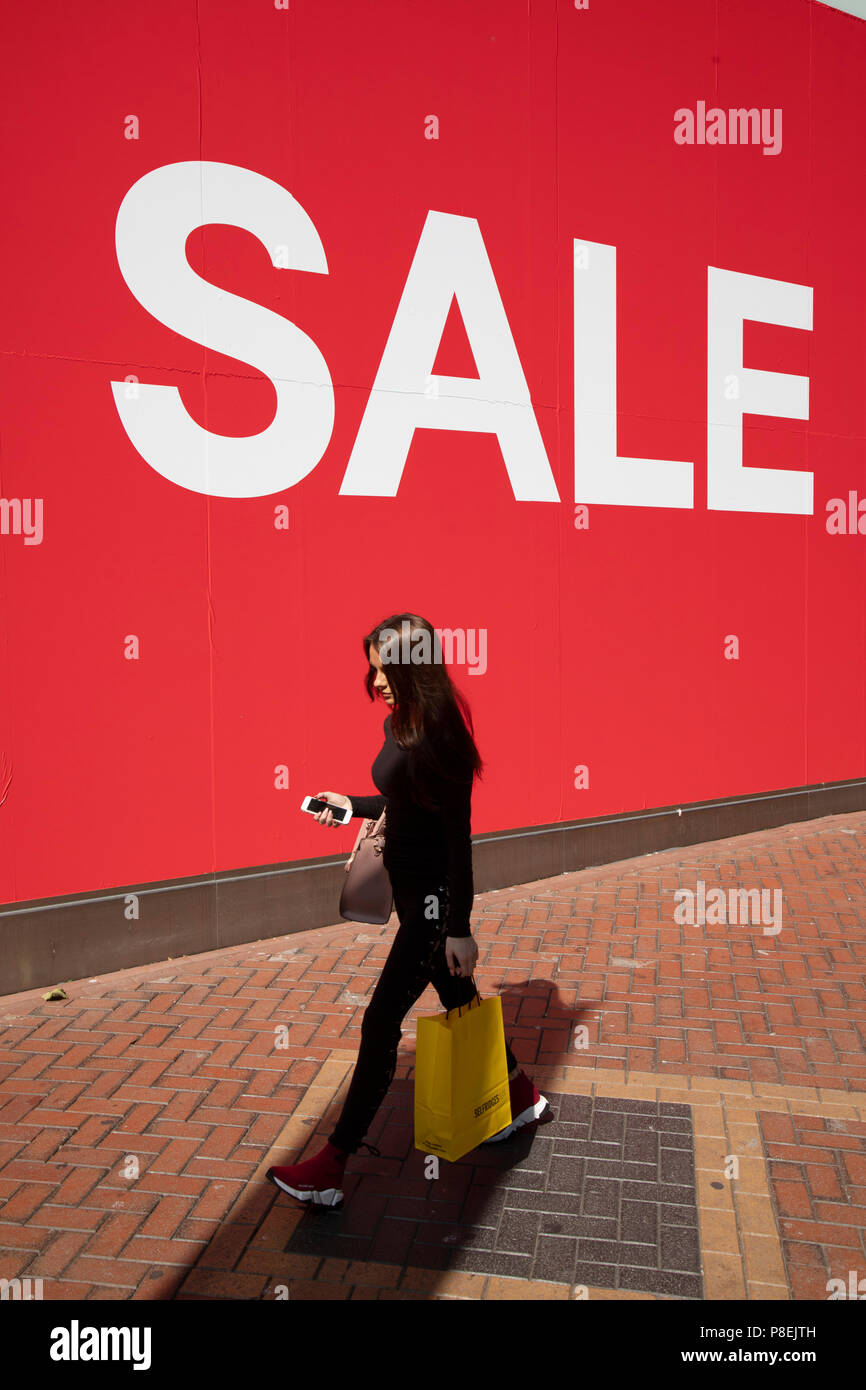 La gente fuori shoppin a piedi passato una scala grande segno di vendita in caratteri bianchi su sfondo rosso al di fuori di H&M, un grande high street abbigliamento negozio di vendita al dettaglio a Birmingham, Regno Unito. Il suo tempo per il summer sales, e la maggior parte dei negozi sono pubblicità di grandi riduzioni dei prezzi. Gli affari sono disponibili e le vie dello shopping sono occupato. Foto Stock