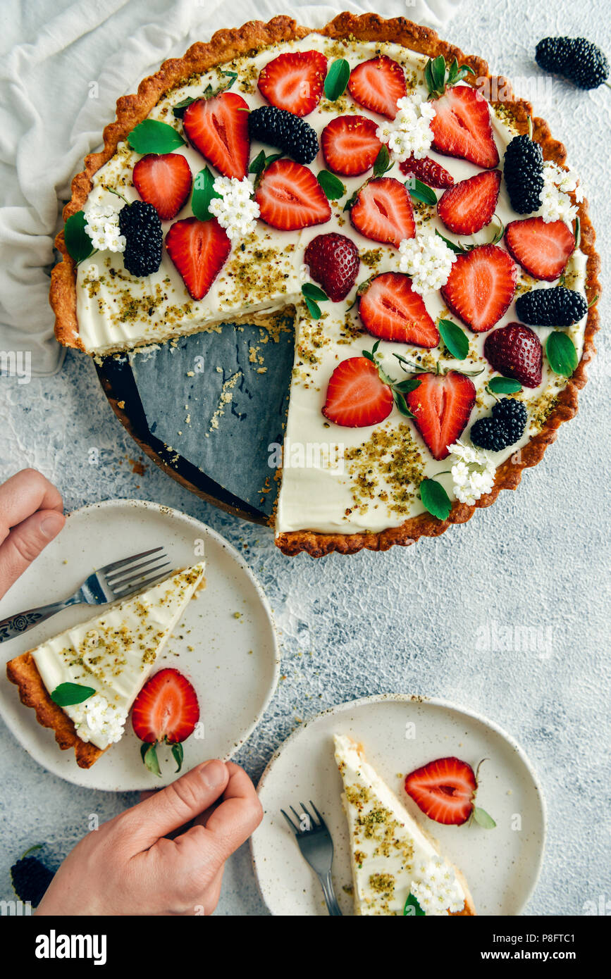 Una donna che serve una fetta di fragola torta di formaggio su una piastra bianca. Un altro strato e il resto della torta di accompagnare con un sacco di fragole e mulbe Foto Stock
