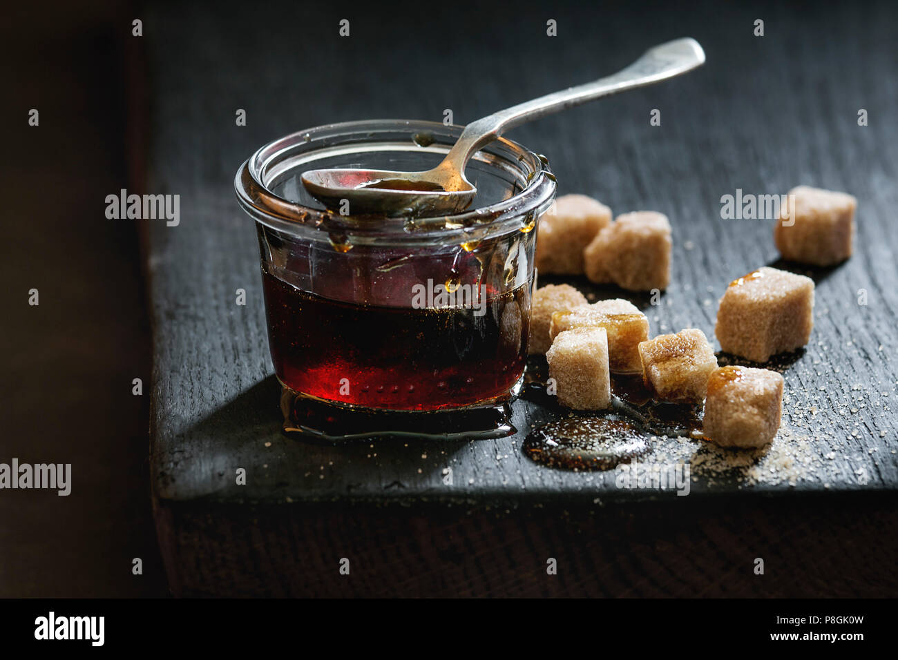 In casa liquido trasparente marrone zucchero caramellato in un barattolo di vetro in piedi sul legno nero con bordo cucchiaio e può zollette di zucchero. Close up. La luce del giorno. Foto Stock