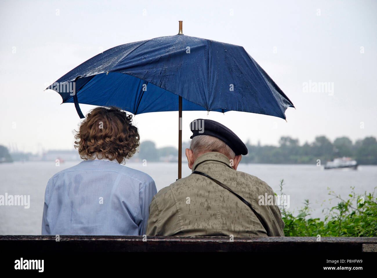 Ein Senioren-Paar auf einer Parkbank an der Promenade in Hamburg-Teufelsbrueck schuetzt sich mit einem Regenschirm gegen einen Sommer-Regen. (C) Marti Foto Stock