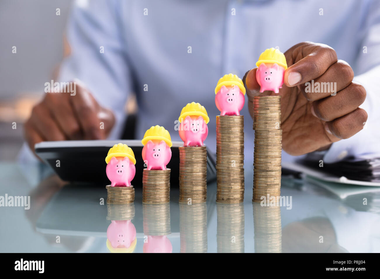 Close-up di persona d'affari immissione Piggybank con Giallo Hardhat sulle monete impilate su scrivania riflettente Foto Stock