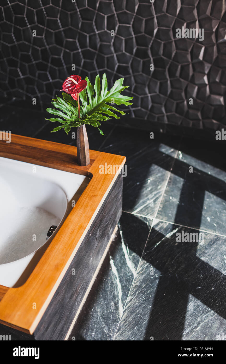 Il design moderno del bagno con textured piastrelle nere e le pareti in legno, bordo della vasca da bagno. La luce dalla finestra e vaso di fiori Foto Stock