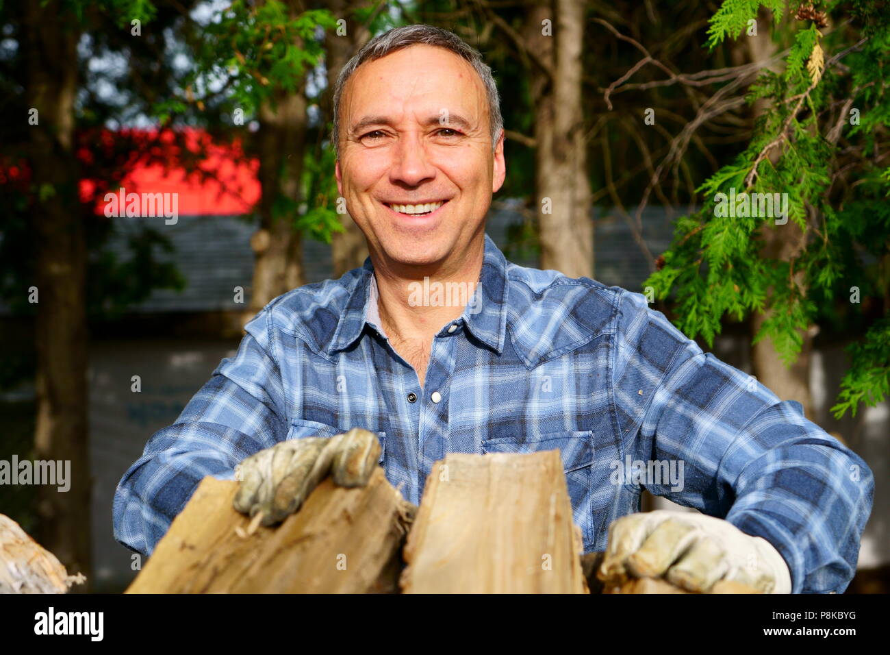 A cinquanta anni uomo caucasico indossando una maglietta bleu è sorridente a noi mentre il suo palificazione in legno per la stagione invernale, al fine di riscaldare la casa. Foto Stock