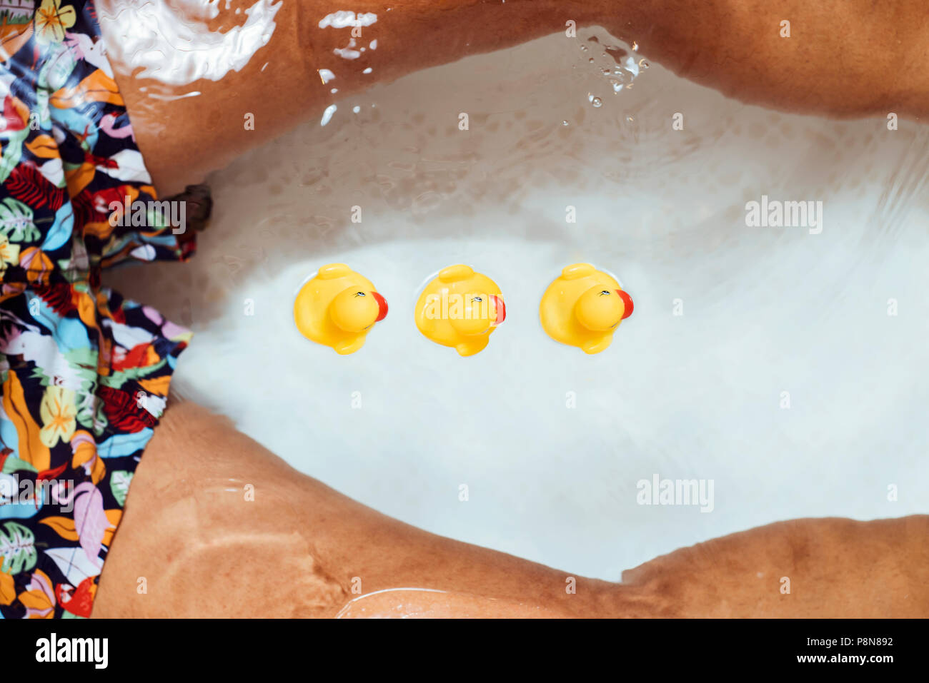 Elevato angolo di visione di un giovane uomo caucasico che indossa un costume da bagno rilassante, circondato da gomma anatre, nella vasca del bagno Foto Stock