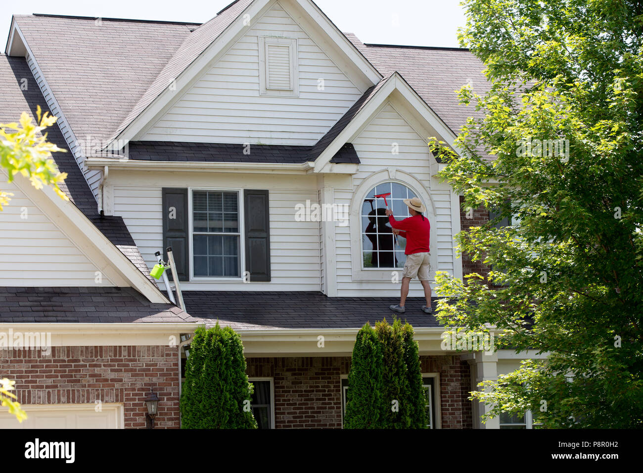 Casa orgoglioso uomo pulizia del piano superiore di windows con un tergipavimento come egli si trova sul patio tetto di una sistemazione di home Foto Stock