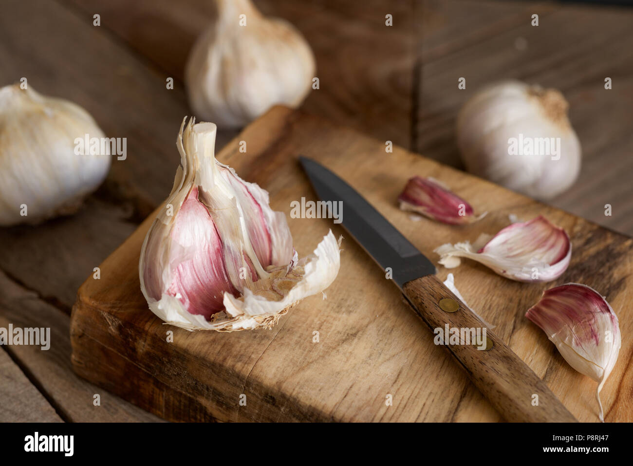 Viola organico bulbi di aglio e chiodi di garofano con una impugnatura in legno coltello seghettato essendo preparato su un tagliere di legno Foto Stock