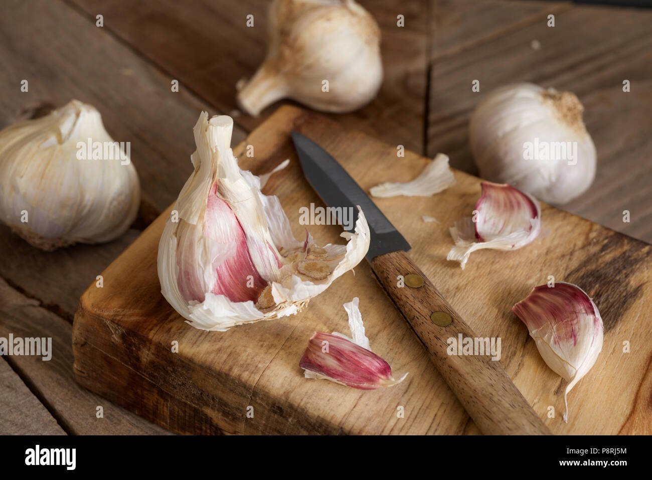 Viola organico bulbi di aglio e chiodi di garofano con una impugnatura in legno coltello seghettato essendo preparato su un tagliere di legno Foto Stock
