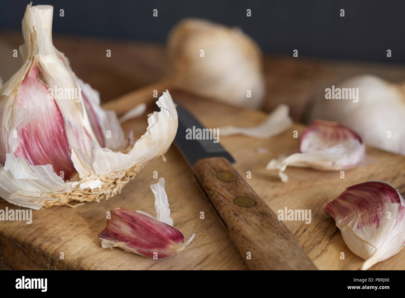 Viola organico bulbi di aglio e chiodi di garofano con una impugnatura in legno coltello seghettato essendo preparato su un tagliere di legno Foto Stock