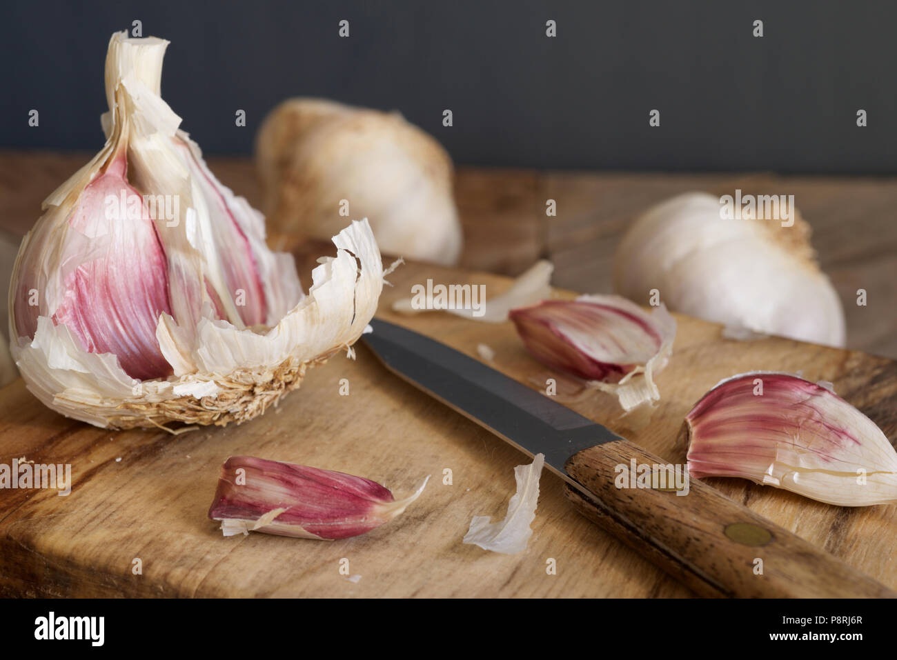 Viola organico bulbi di aglio e chiodi di garofano con una impugnatura in legno coltello seghettato essendo preparato su un tagliere di legno Foto Stock