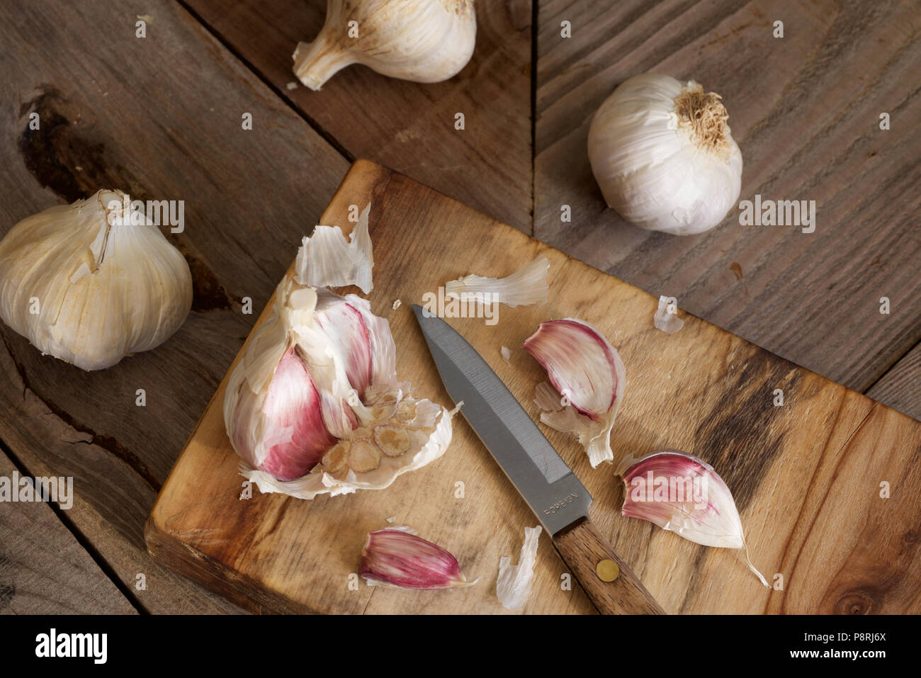 Viola organico bulbi di aglio e chiodi di garofano con una impugnatura in legno coltello seghettato essendo preparato su un tagliere di legno Foto Stock