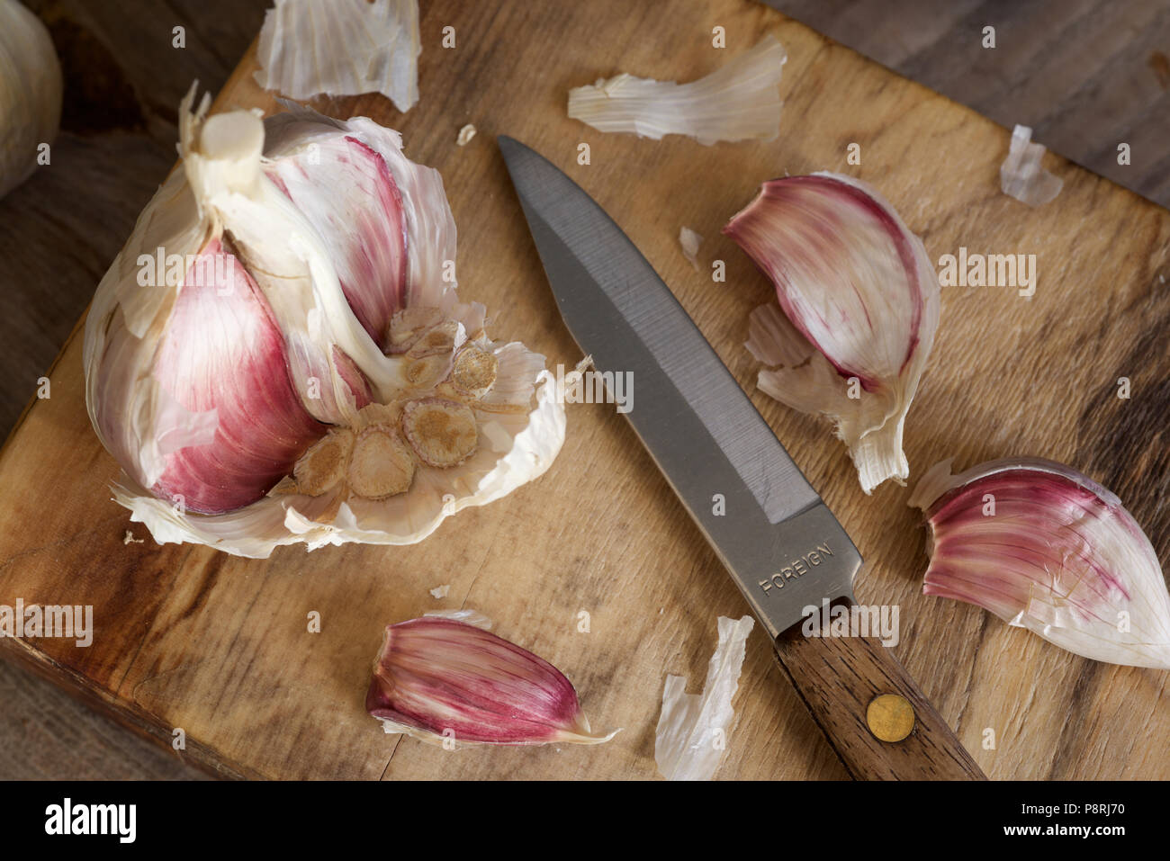 Viola organico bulbi di aglio e chiodi di garofano con una impugnatura in legno coltello seghettato essendo preparato su un tagliere di legno Foto Stock